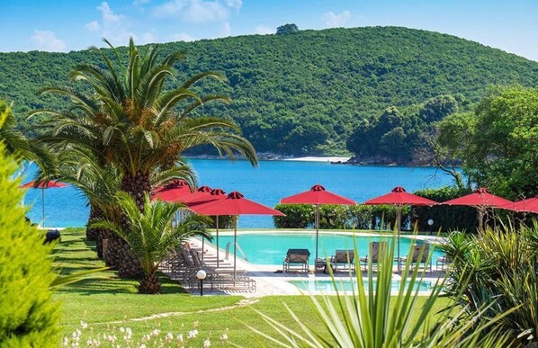 A pool area with red umbrellas, lounge chairs, and a view of the sea and forested hills. - Bella Mare Hotel (Corfu)