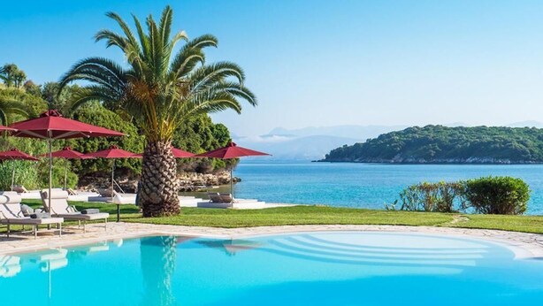 A pool with lounge chairs and umbrellas, a palm tree, and a view of the sea and mountains. - Bella Mare Hotel (Corfu)