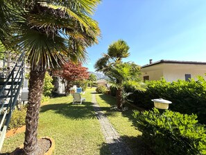 A well-maintained garden with a pathway leading to a white building, surrounded by various plants and a palm tree. - Apartment House Ammura Balcony Garden (Tignale)