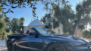 En svart cabriolet står parkerad på en grusuppfart, med träd och en byggnad i bakgrunden.