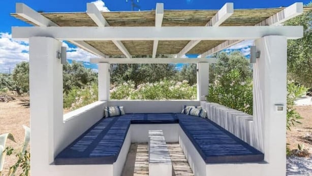 Ett takförsett utomhusområde med sittplatser, en vit pergola, inbyggda bänkar och utsikt över det omgivande landskapet.