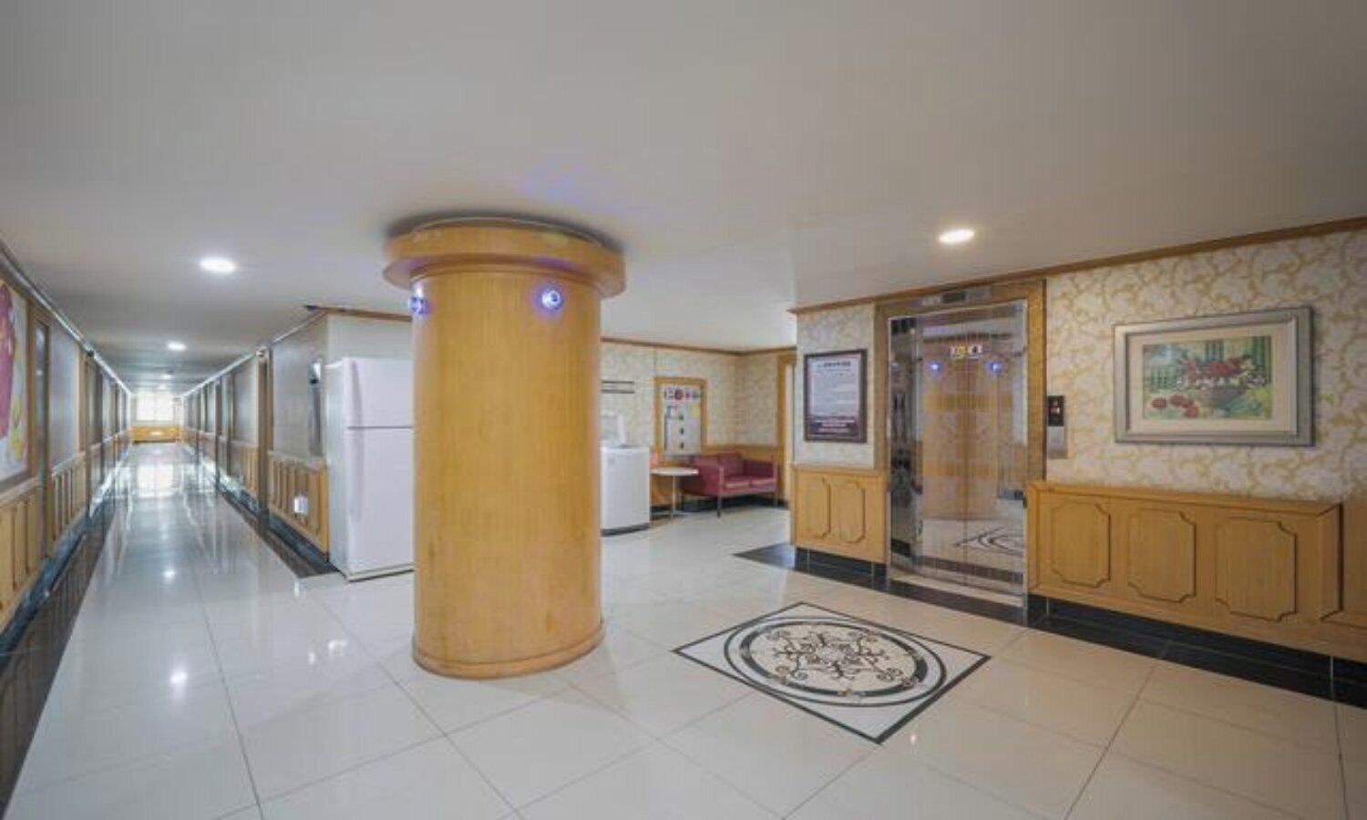 Pasillo de hotel con una columna central, diseño de piso estampado y una zona de recepción con mostrador y asientos.