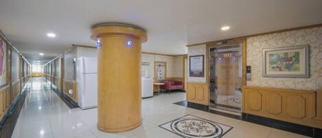Pasillo de hotel con una columna central, diseño de piso estampado y una zona de recepción con mostrador y asientos.