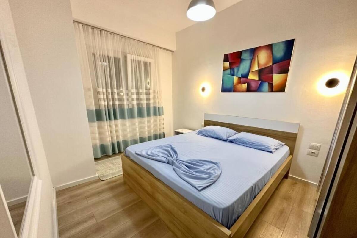 A hotel room with a wooden bed, a large window with sheer curtains, a colorful abstract painting on the wall, and a ceiling light.