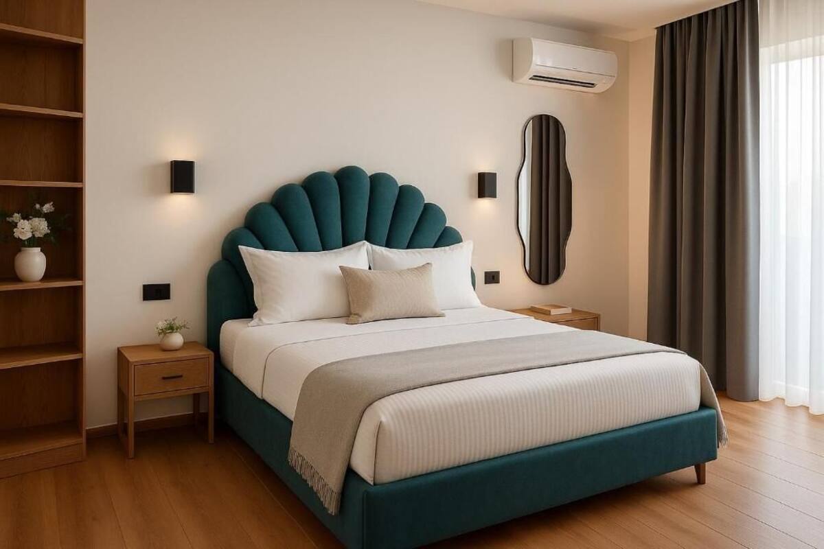 A modern bedroom with a teal upholstered headboard, a white bedspread, a bedside table with a vase, and an air conditioning unit on the wall.