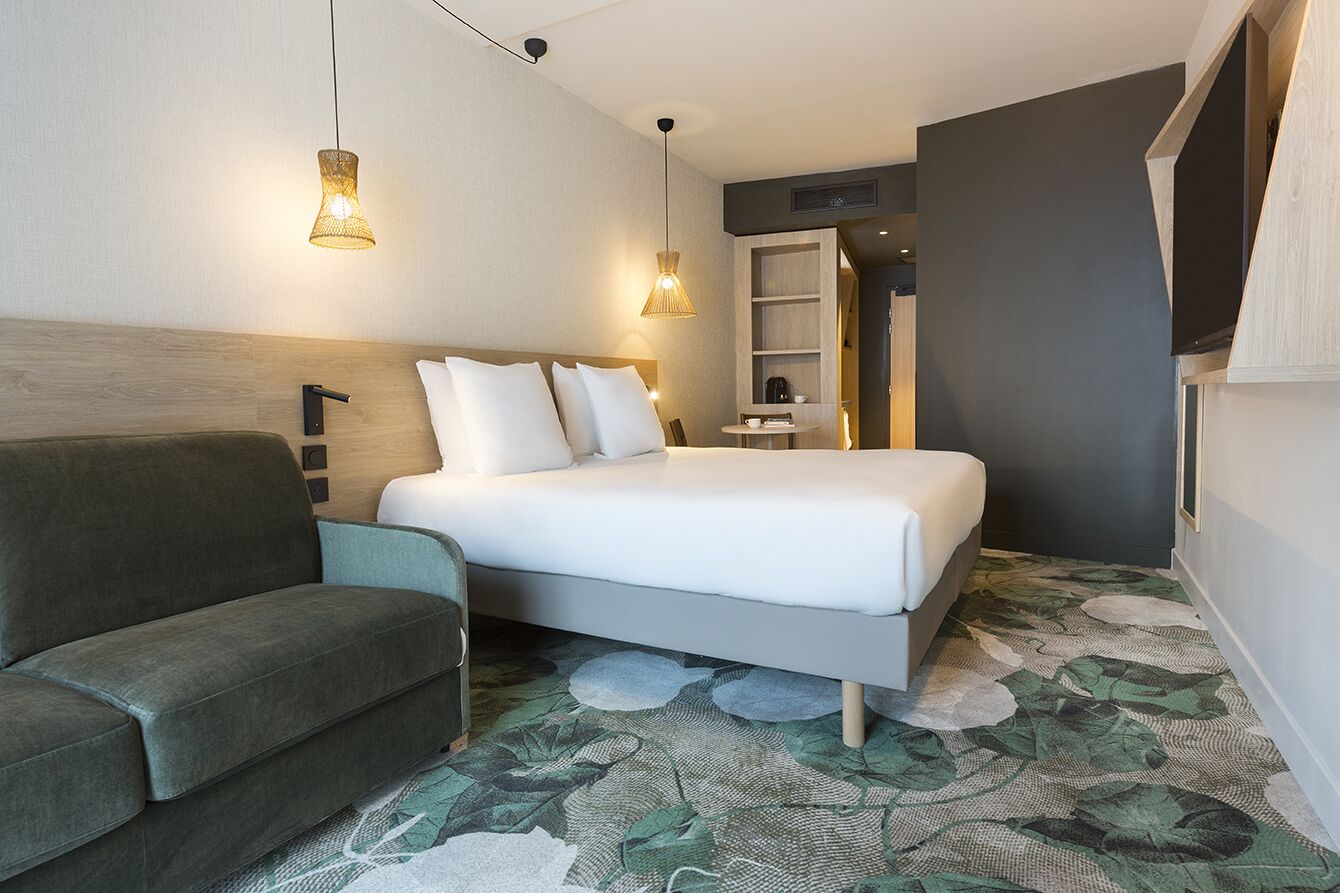 A modern hotel room with a bed, a sofa, a small table, and a bookshelf.