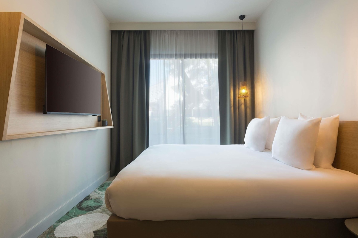 A hotel room with a bed, a flat-screen TV mounted on the wall, a window with curtains, and a wall-mounted lamp.