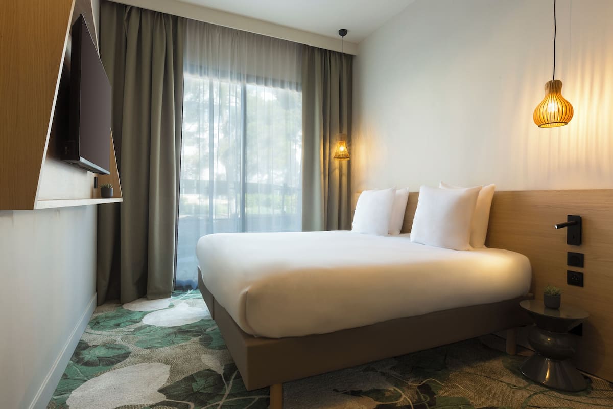 A hotel room with a bed, a TV, a small table with a plant, a wall-mounted light, and a window with curtains.
