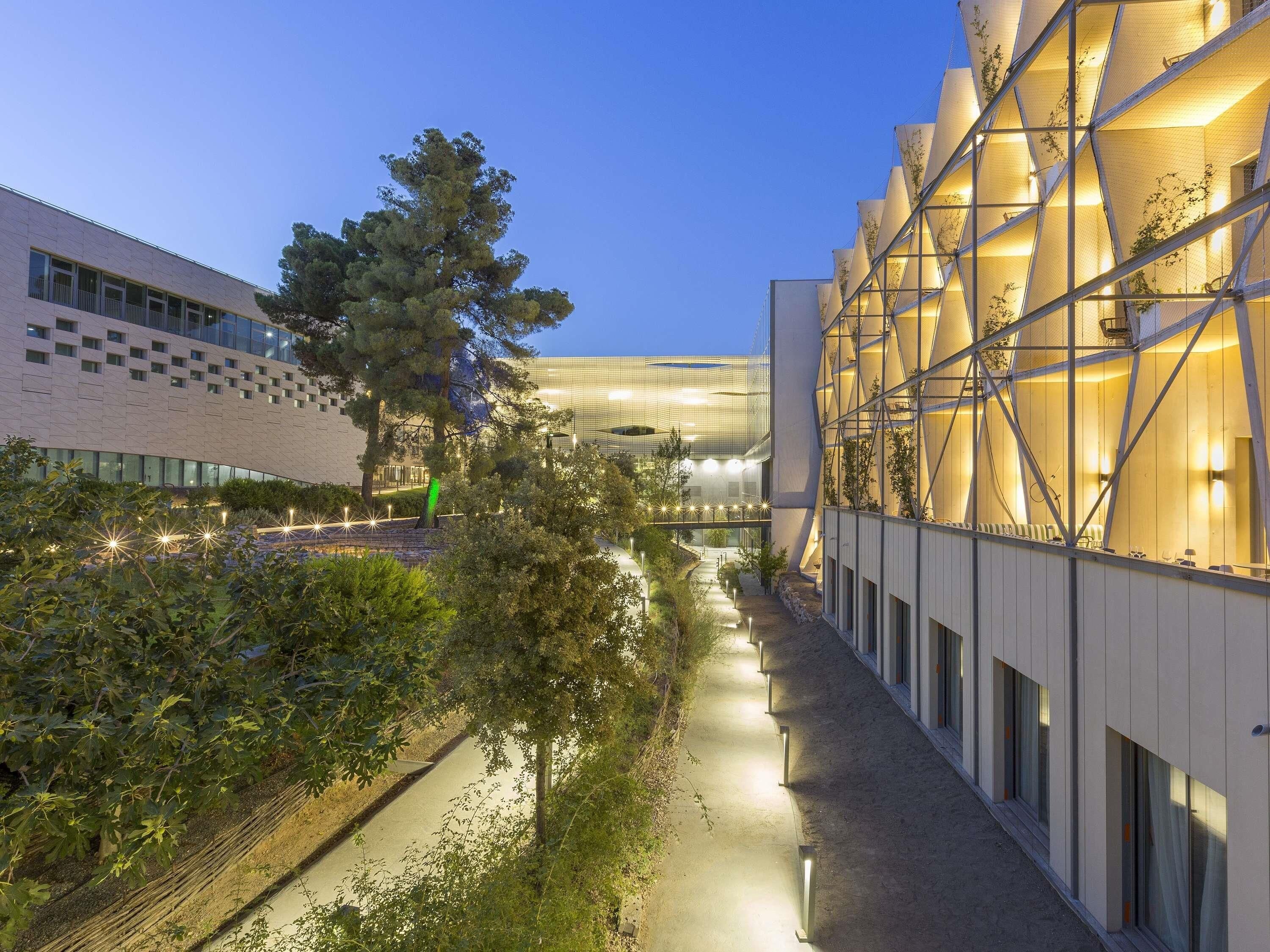 A modern building with a geometric facade, a pathway lined with trees, and a clear sky.