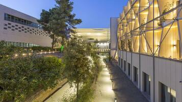 A modern building with a geometric facade, a pathway lined with trees, and a clear sky.