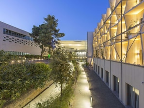 A modern building with a geometric facade, a pathway lined with trees, and a clear sky.