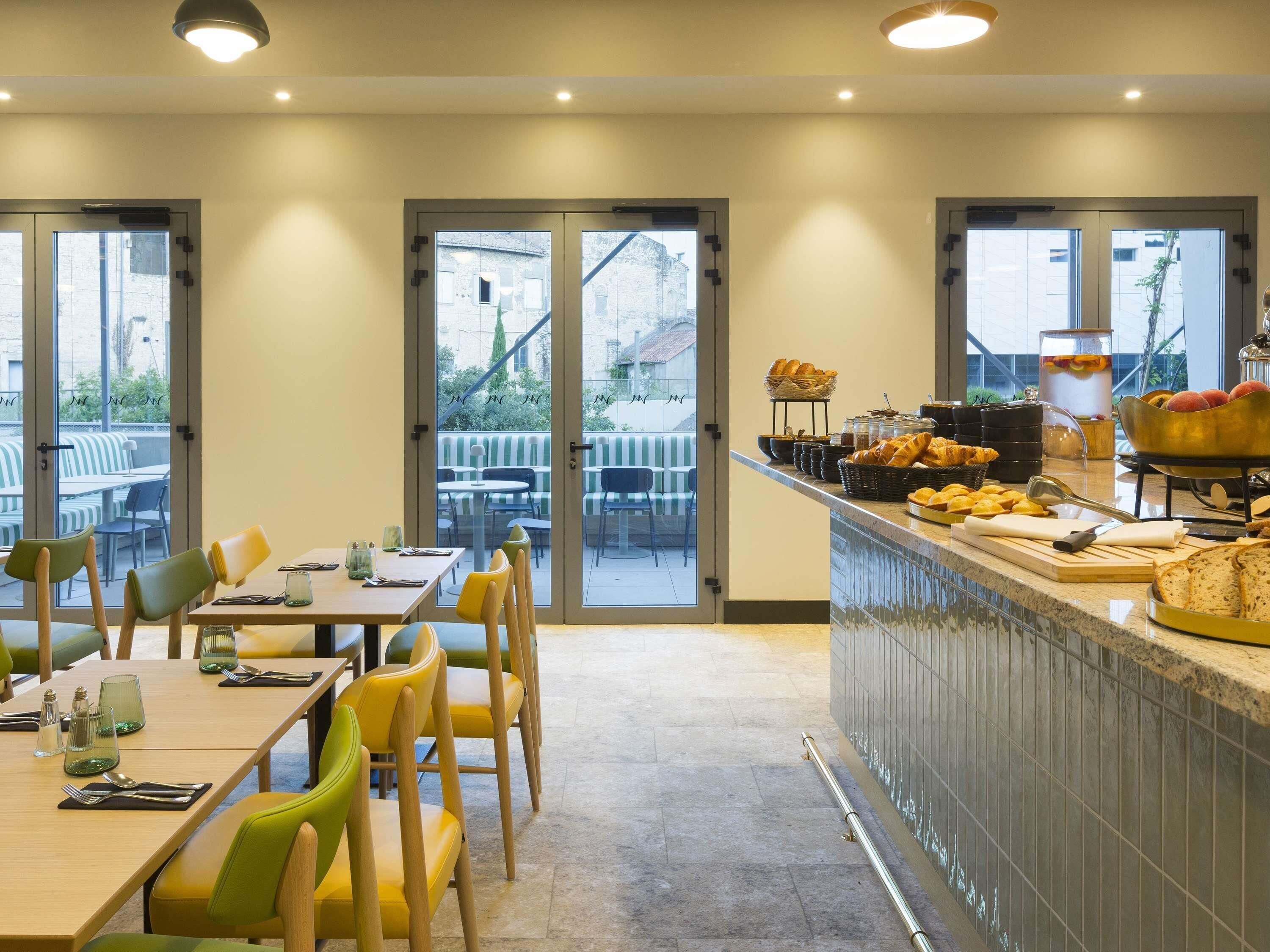 A restaurant with a counter displaying bread and pastries, a dining area with tables and chairs, and large windows offering a view of the outside.
