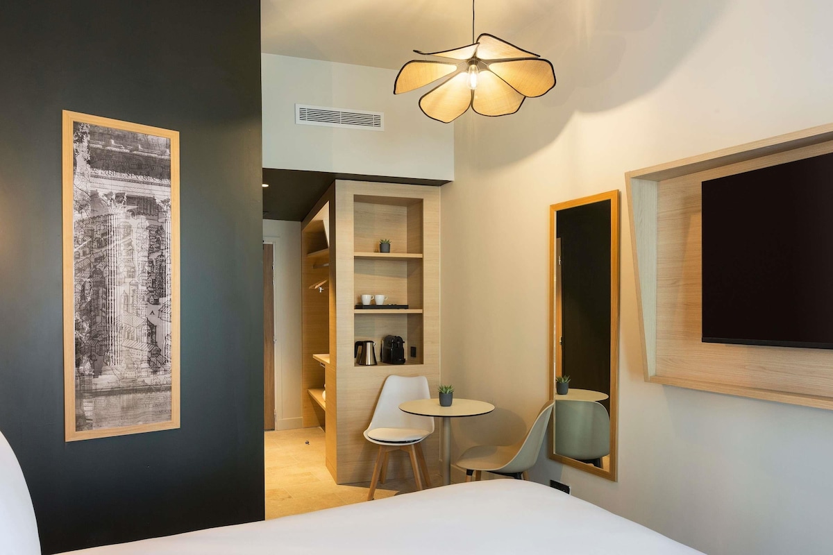 A modern hotel room with a bed, a desk, a chair, a small table, and a large mirror. There is a framed picture on the wall and a ceiling light fixture.