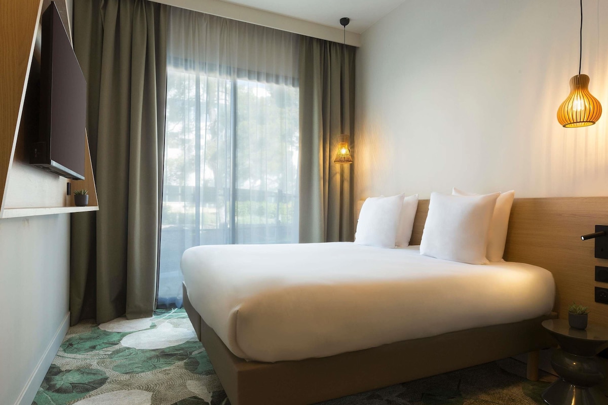 A hotel room with a bed, curtains, a wall-mounted TV, a bedside lamp, and a small table with a plant.