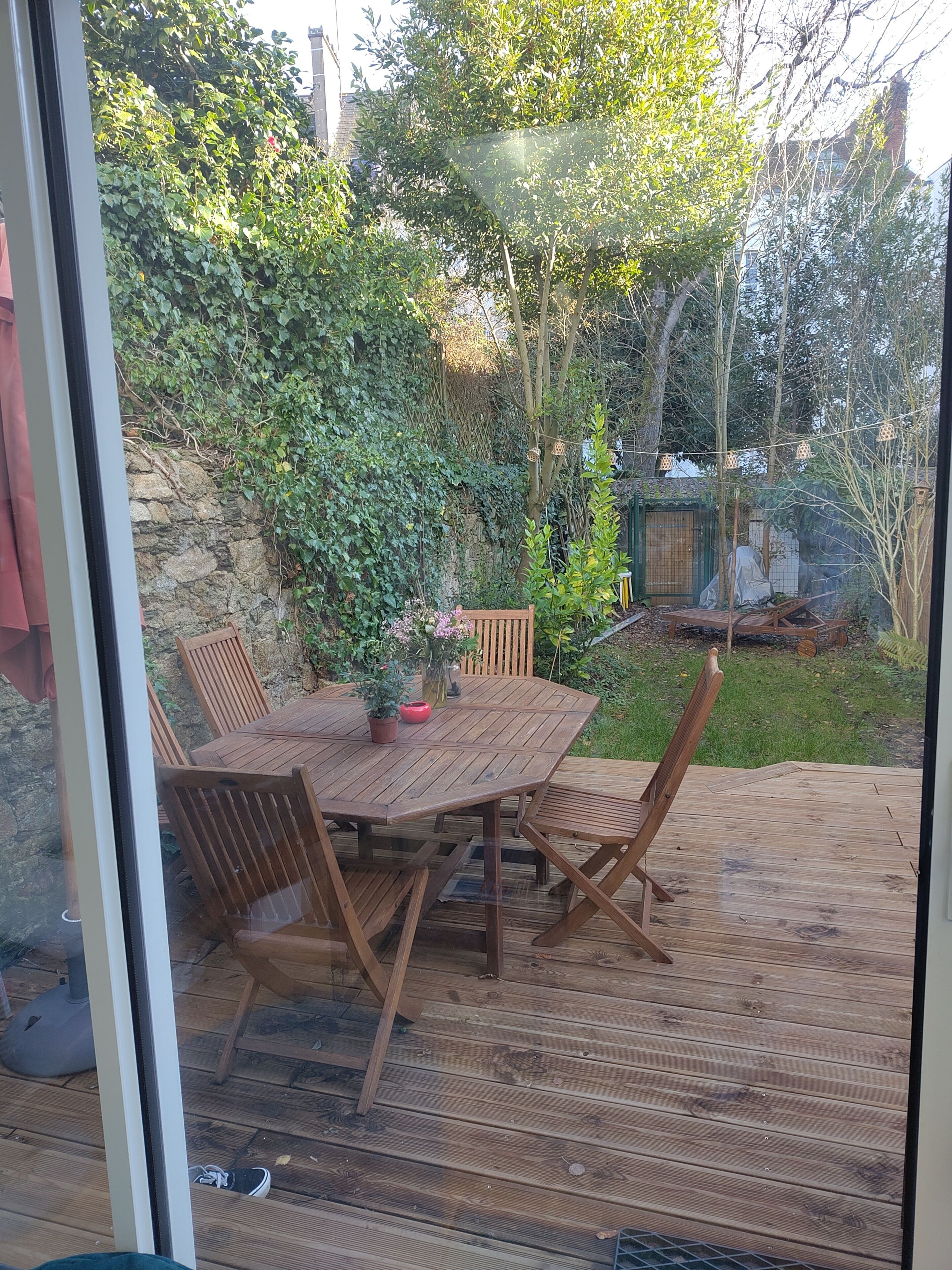 En träveranda med ett runt bord och stolar, omgiven av grönska och en stenmur.