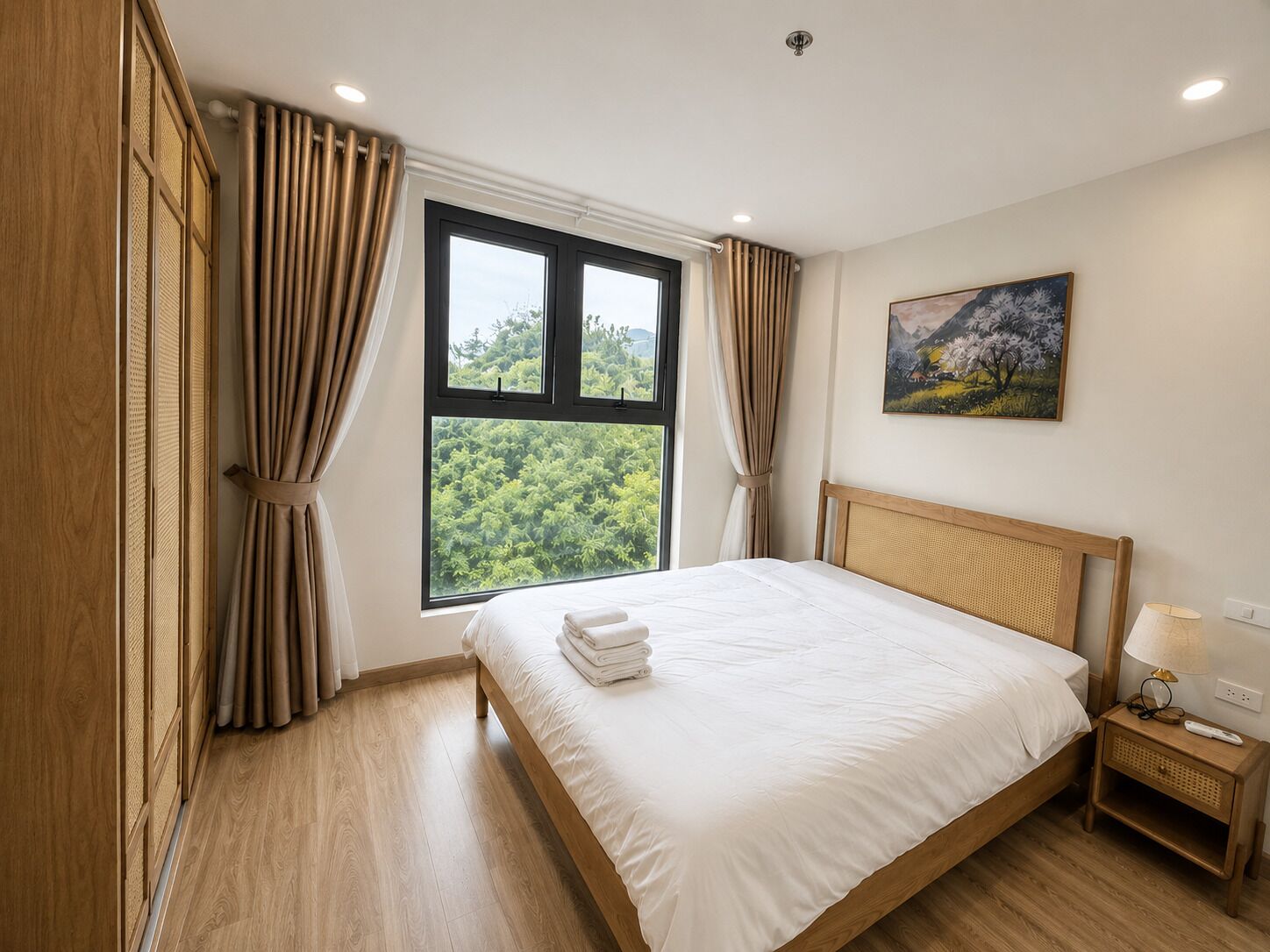 A bedroom with a large window, a bed, a nightstand, and a painting on the wall.