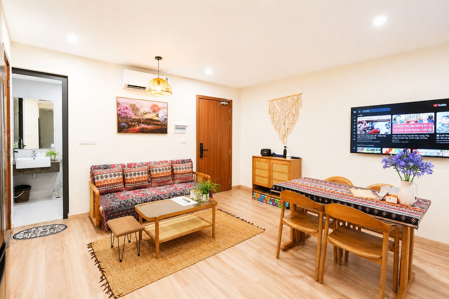 Un salon comprenant un canapé, une table basse, une table à manger et des chaises. Un téléviseur est fixé au mur, ainsi qu’un meuble en bois et une porte donnant sur une autre pièce.