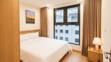 A hotel room with a bed, a nightstand with a lamp, a window with curtains, and a painting on the wall.