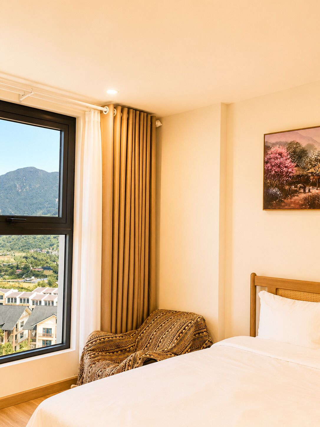 A hotel room with a bed, a window with curtains, a painting on the wall, and a view of buildings and hills.