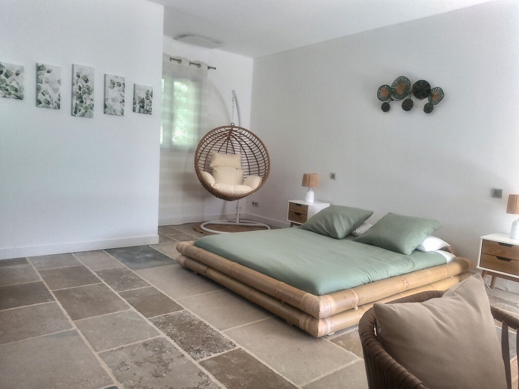 A bedroom with a bed, a hanging chair, a nightstand, and a wall with artwork.