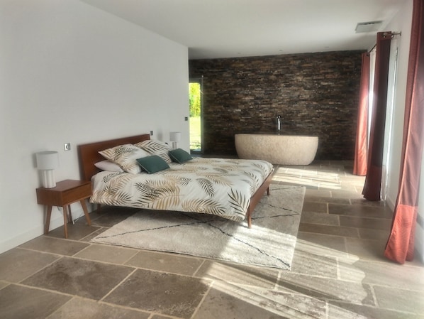 Ein Schlafzimmer mit Bett, Nachttisch, großem Fenster, Steinwand und Badewanne.