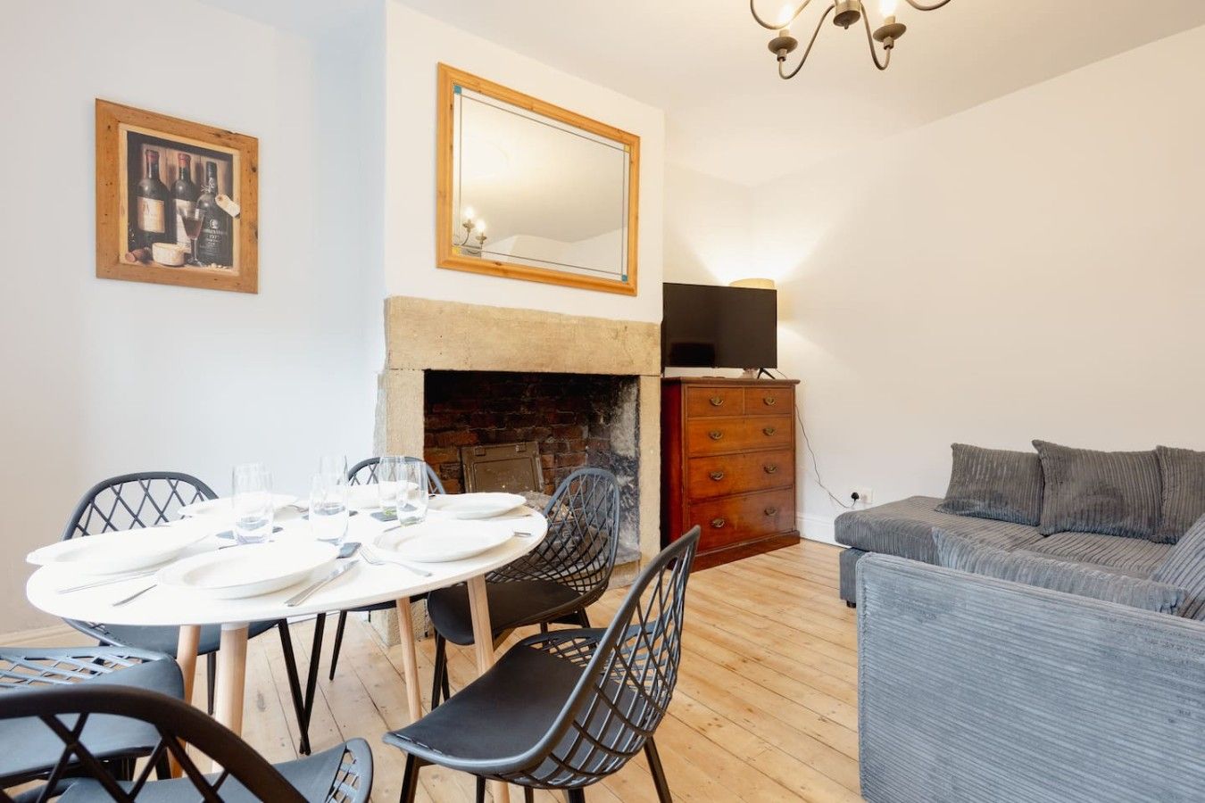 A dining area with a round table set for four, a fireplace, a television on a dresser, and a grey sofa.