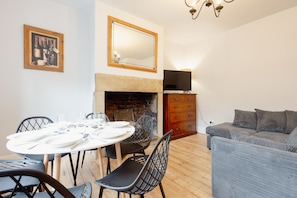 A dining area with a round table set for four, a fireplace, a television on a wooden cabinet, and a grey sofa.