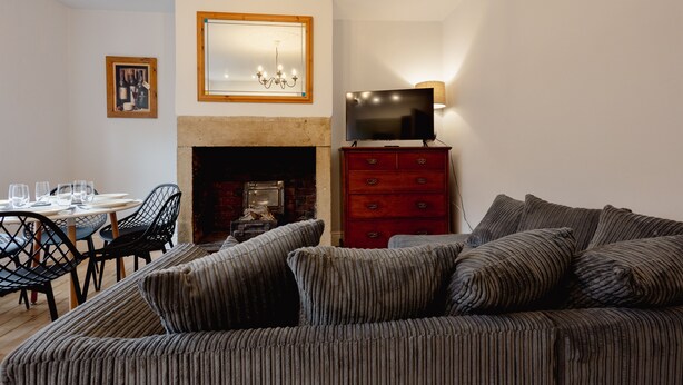 A living room with a fireplace, a television on a red dresser, a sofa, and a dining area with chairs and a table. - Central Hebden Bridge Cottage | Sleeps 6 | Walk to Shops, Bars & Cafés (West Yorkshire)