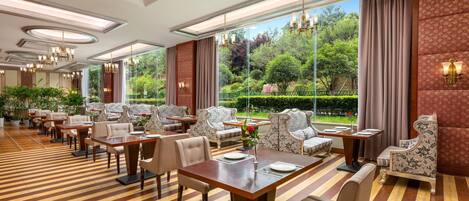 A hotel dining area with multiple tables and chairs, large windows with a view of greenery, and decorative lighting.