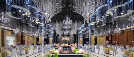 Un salón de banquetes con un escenario central, rodeado de mesas con sillas, y un techo decorado con luces colgantes y candelabros.