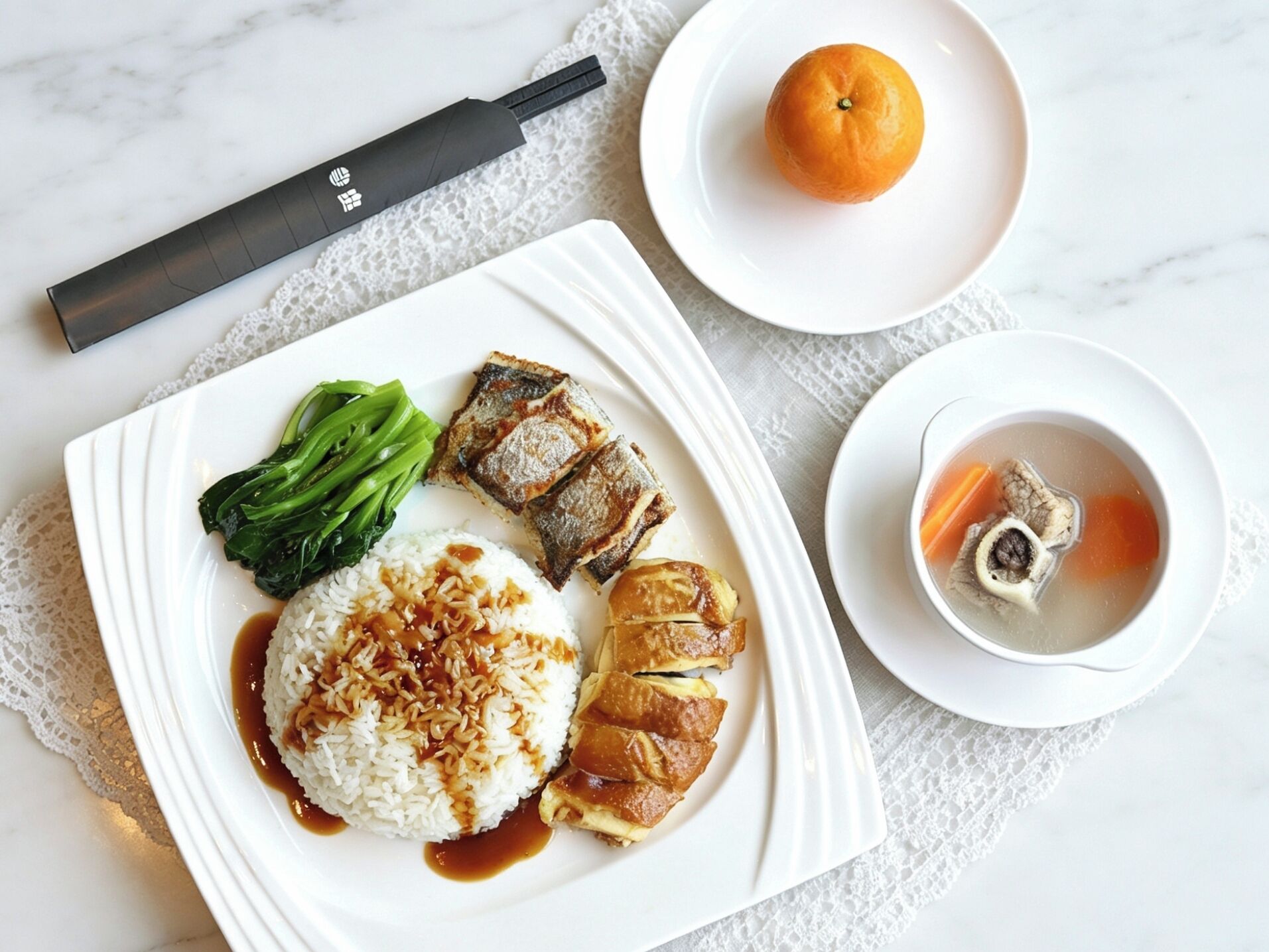 A neatly arranged meal on a white plate with rice, vegetables, and meat, accompanied by a bowl of soup and an orange on a white table.