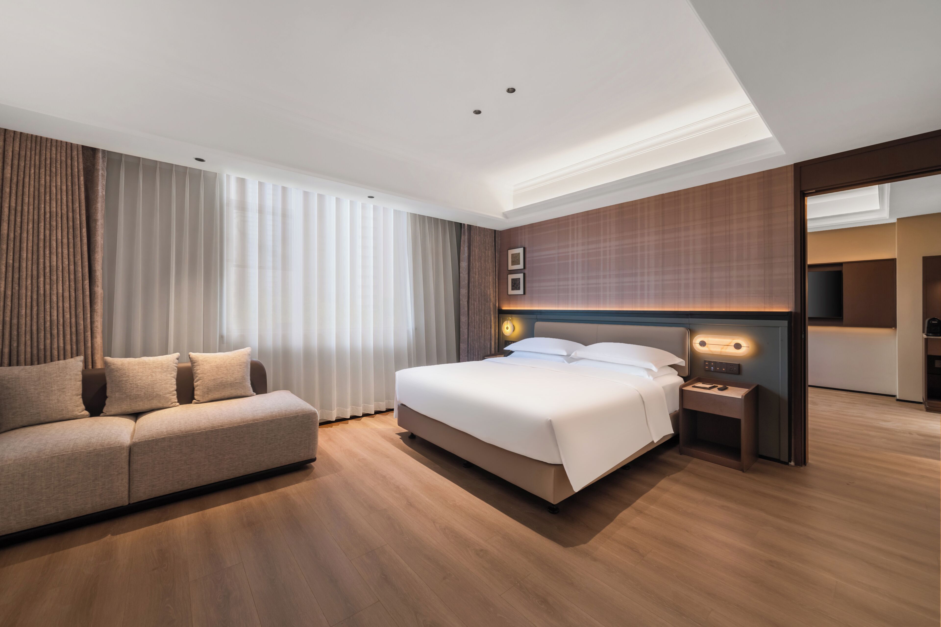A modern hotel room with a large bed, a sofa, a nightstand, and a wall-mounted light.
