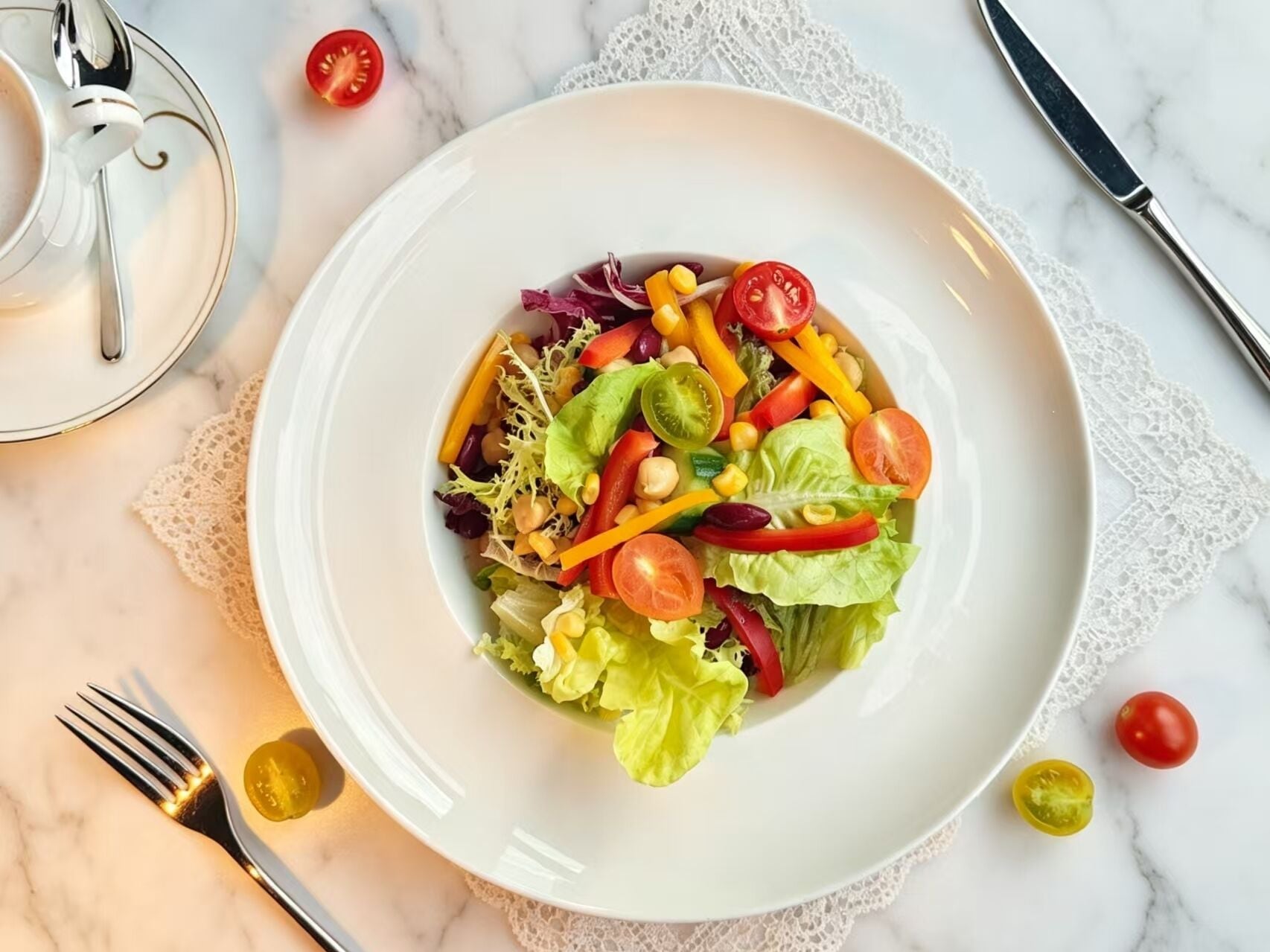 A white plate with a colorful salad, a fork, a knife, and a small glass of yellow liquid on a marble surface.