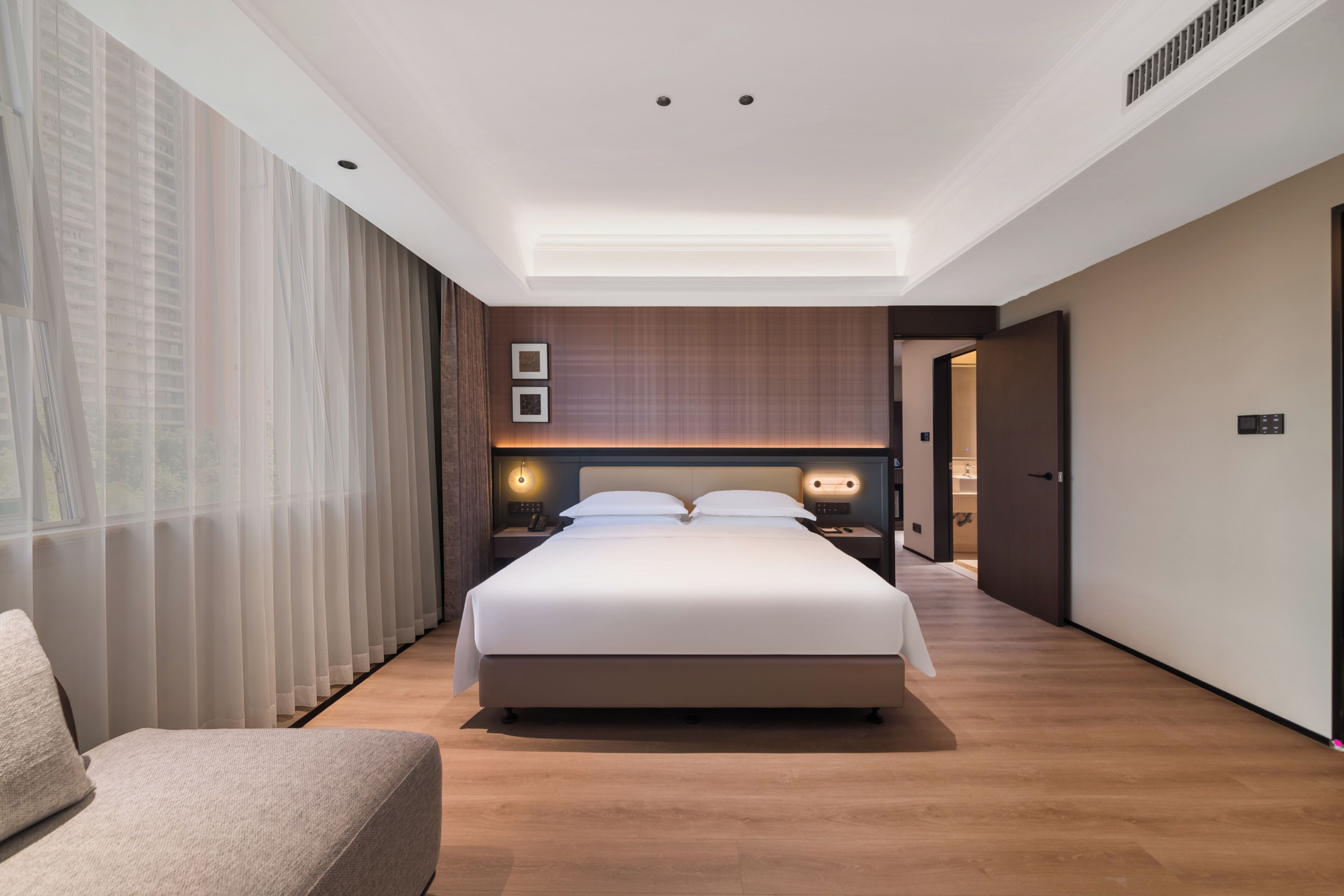 A modern hotel room with a large bed, bedside tables, a chair, and a view of the city through sheer curtains.