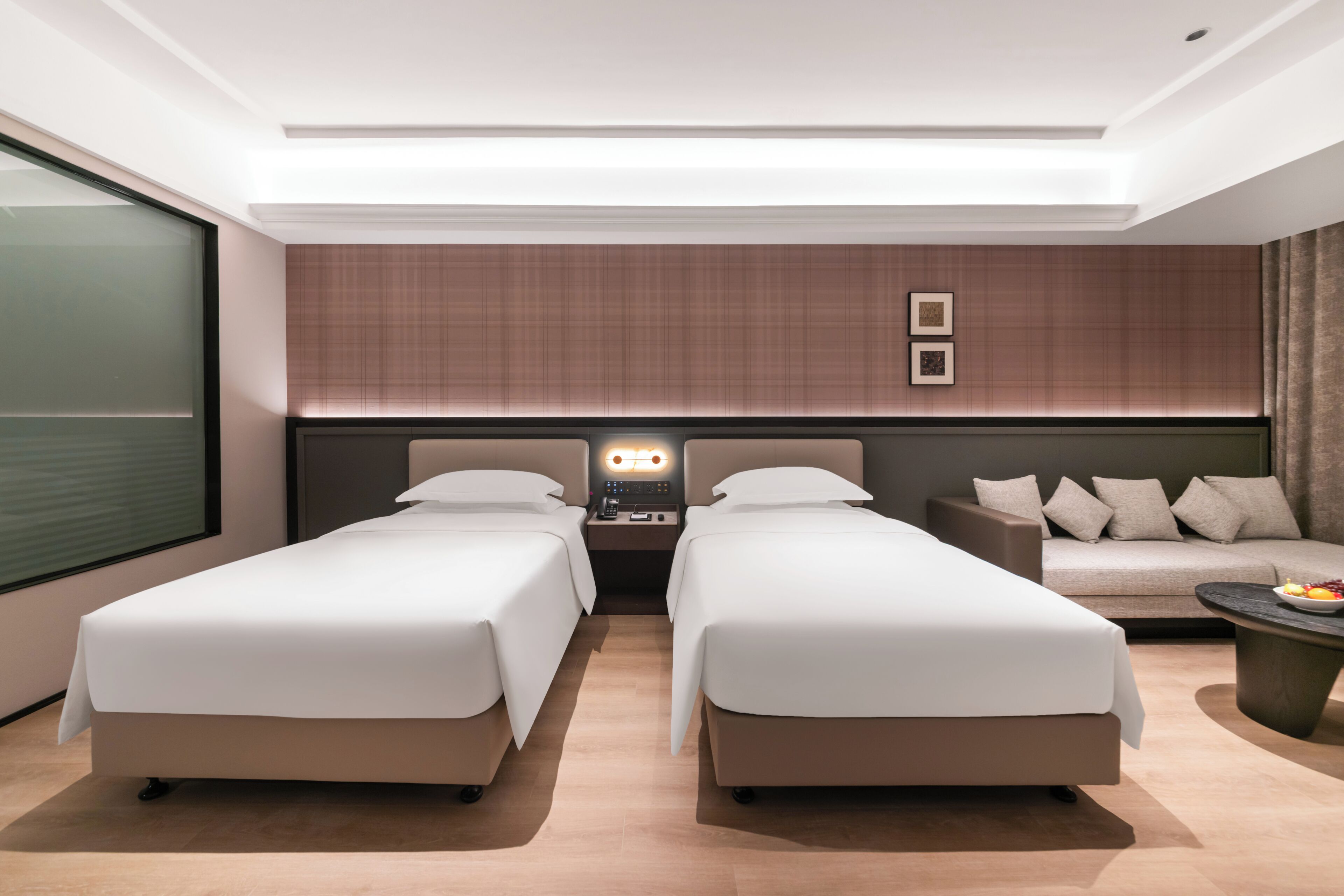 Habitación de hotel con dos camas, un sofá, una mesita de noche con lámpara y vistas al exterior.