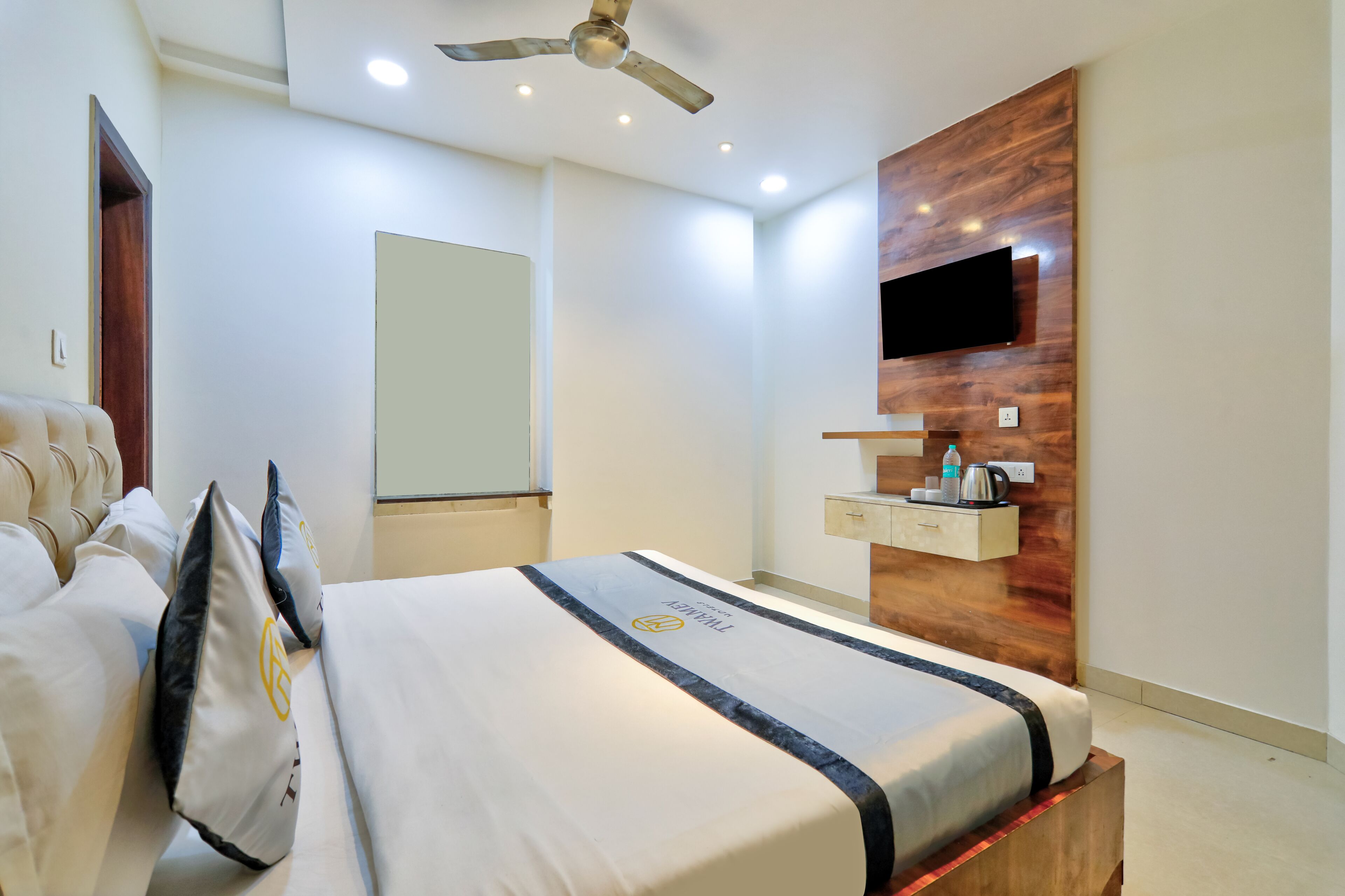 A hotel room with a bed, a ceiling fan, a mounted TV, a wooden accent wall, and a small shelf with a kettle and a bottle.