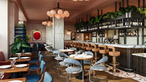 Interior de restaurante con mesas redondas, sillas azules, una zona de bar con estantes llenos de botellas y lámparas esféricas naranjas colgantes.