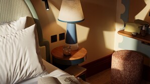 Habitación de hotel con cama, mesita de noche con lámpara, un taburete pequeño y un libro.