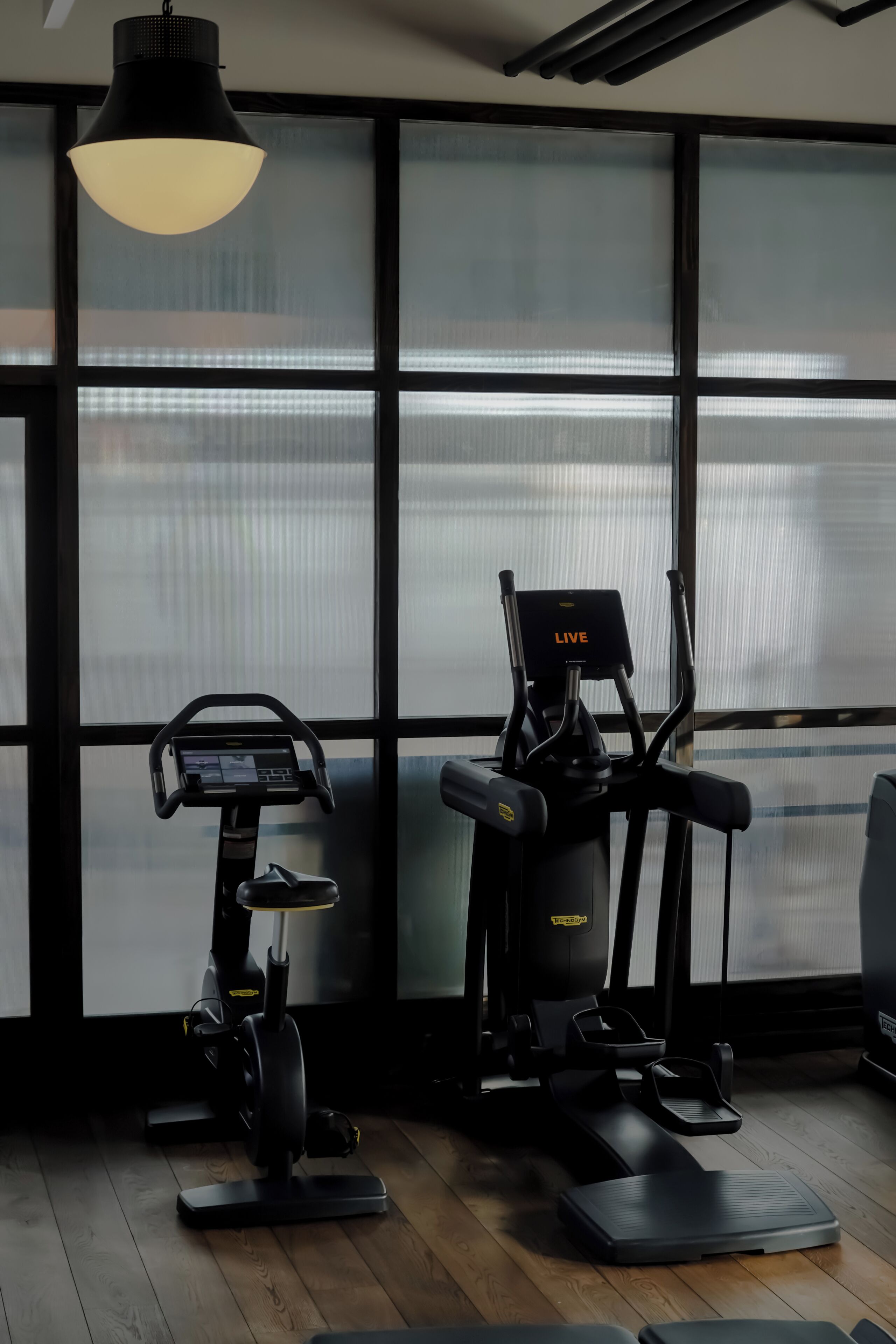 Ginásio interior com equipamentos de exercício, janelas amplas e um candeeiro pendente.