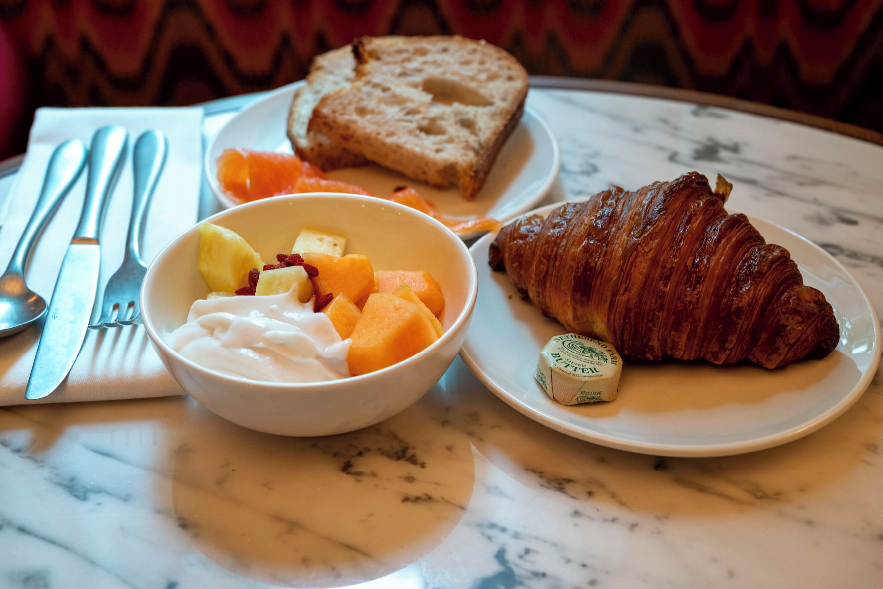 A breakfast setting with a croissant, butter, fruit, and yogurt on a marble table.