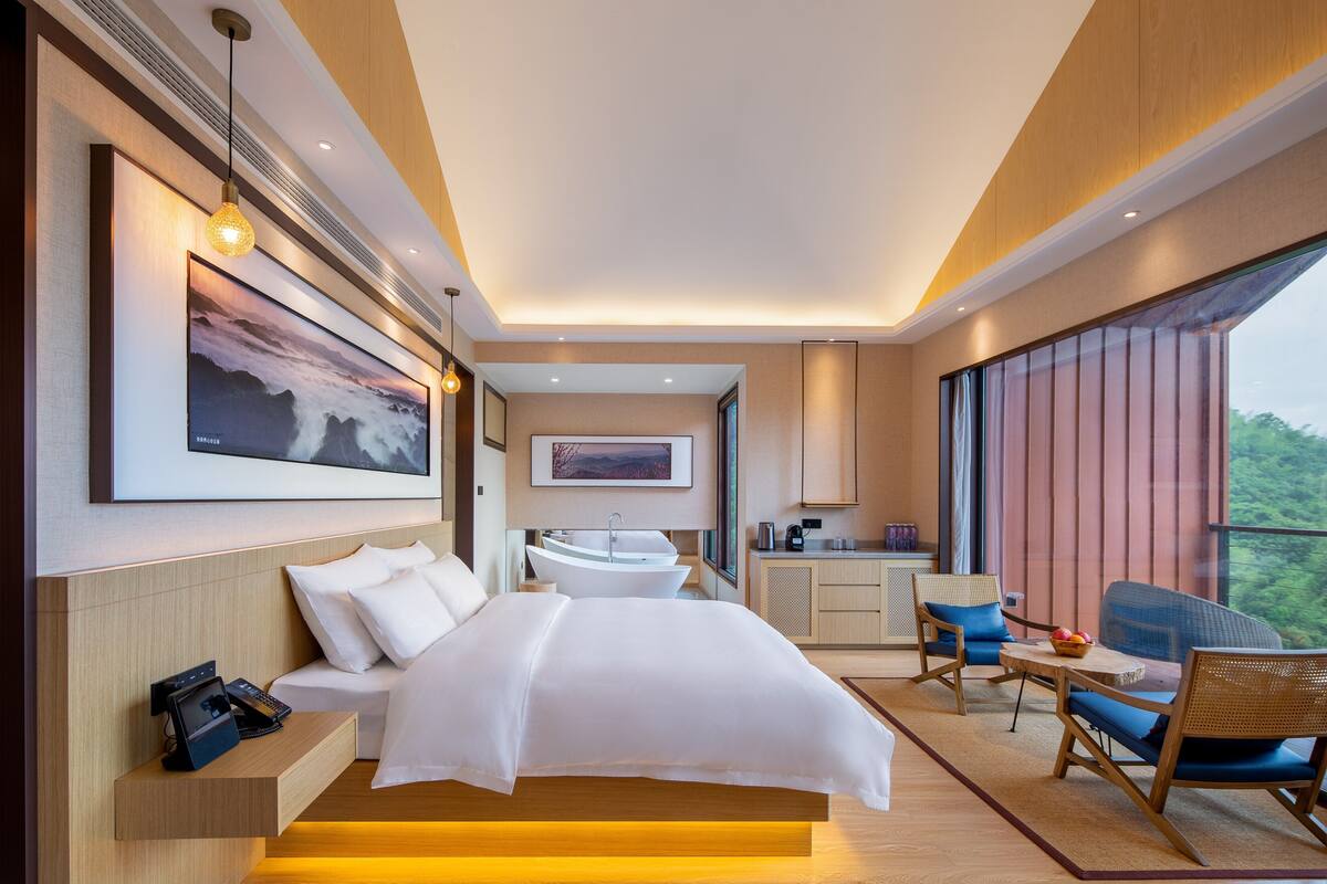 A modern hotel room with a bed, bedside table, phone, artwork, bathroom, and a balcony with seating.