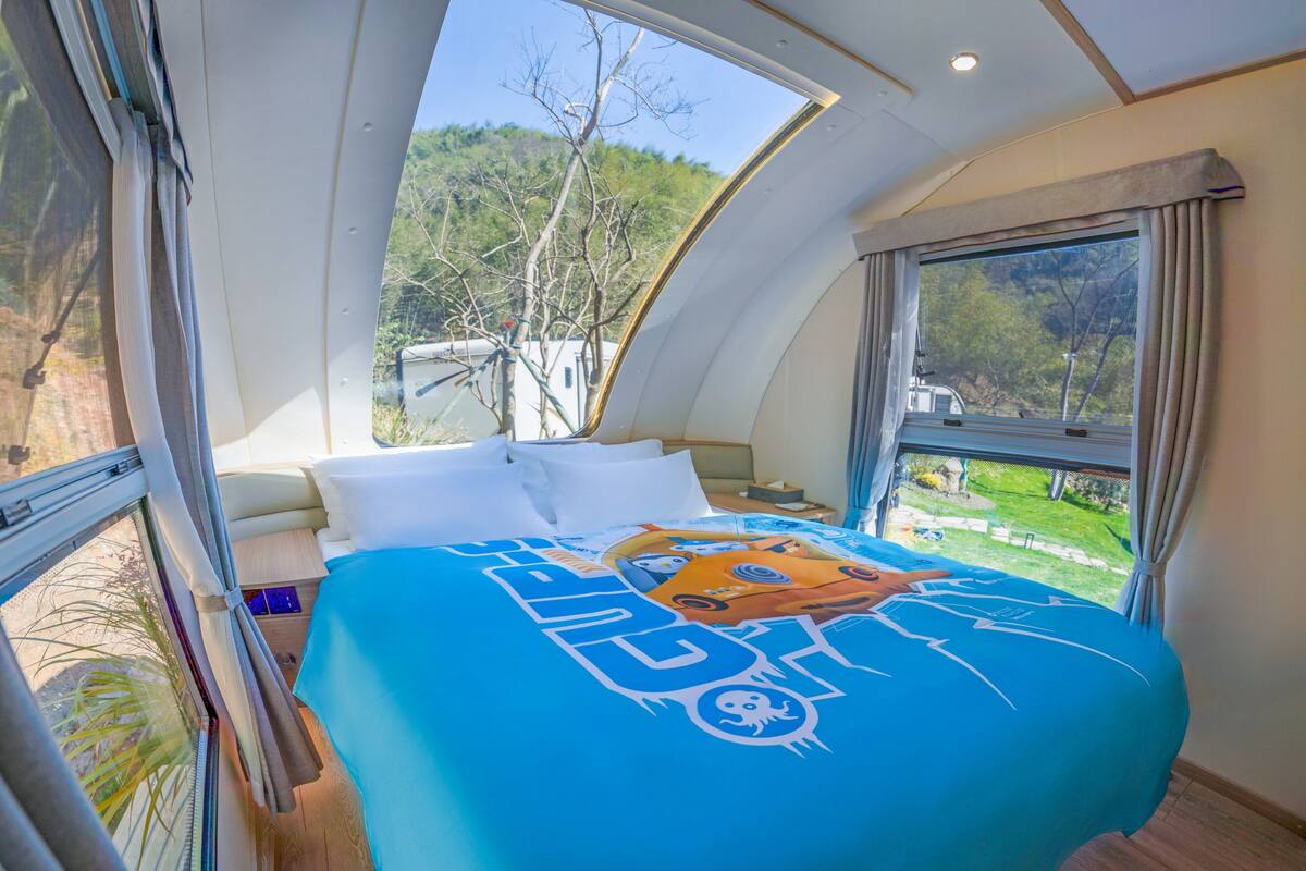 A bedroom with a curved ceiling, a bed with a blue bedspread, and large windows offering a view of the outdoors.