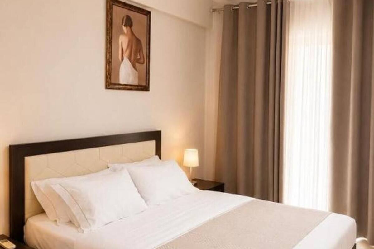 Habitación de hotel con cama, mesita de noche, lámpara, un cuadro en la pared y ventana con cortinas.