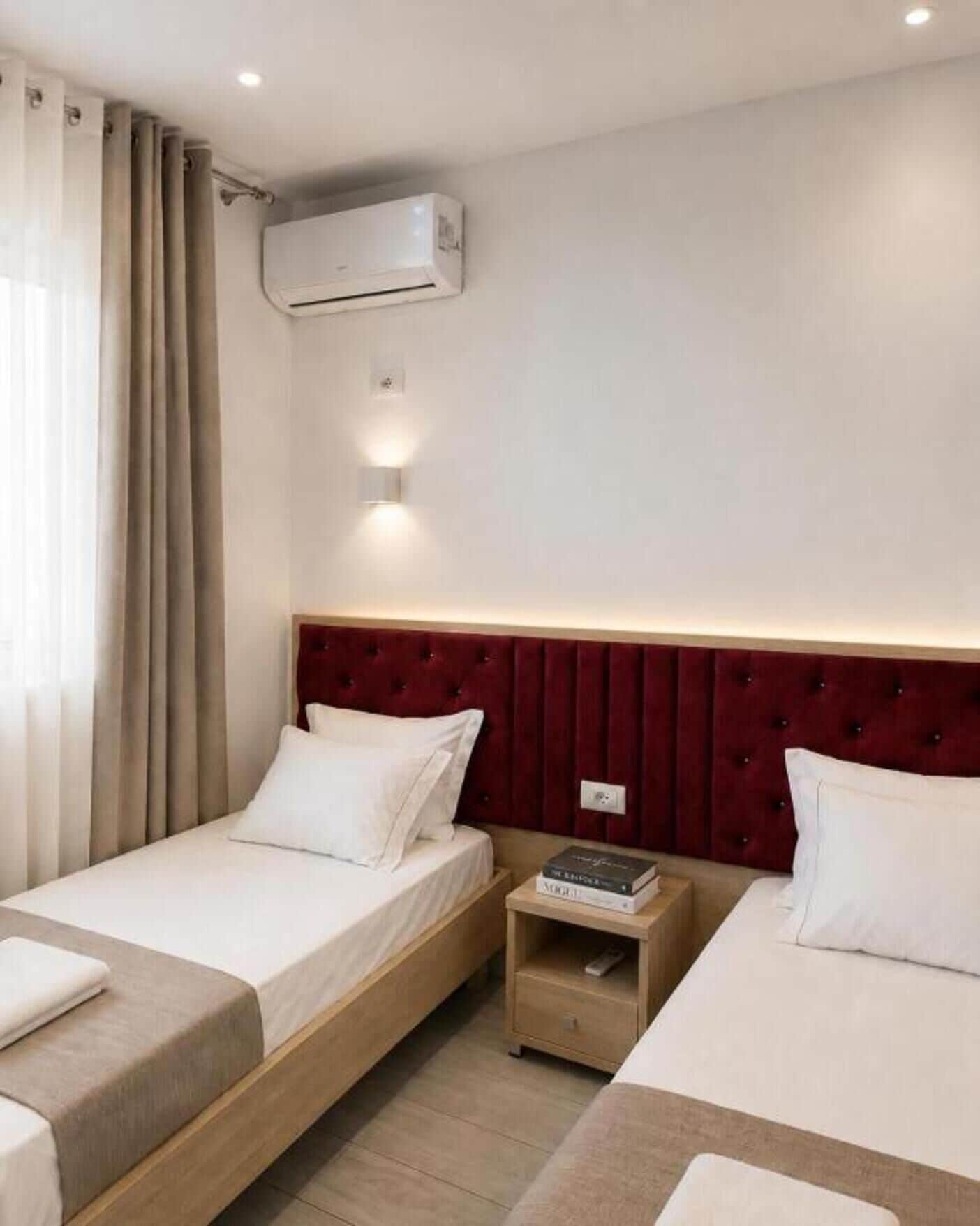 A hotel room with two beds, a red headboard, an air conditioning unit, and a bedside table with books.