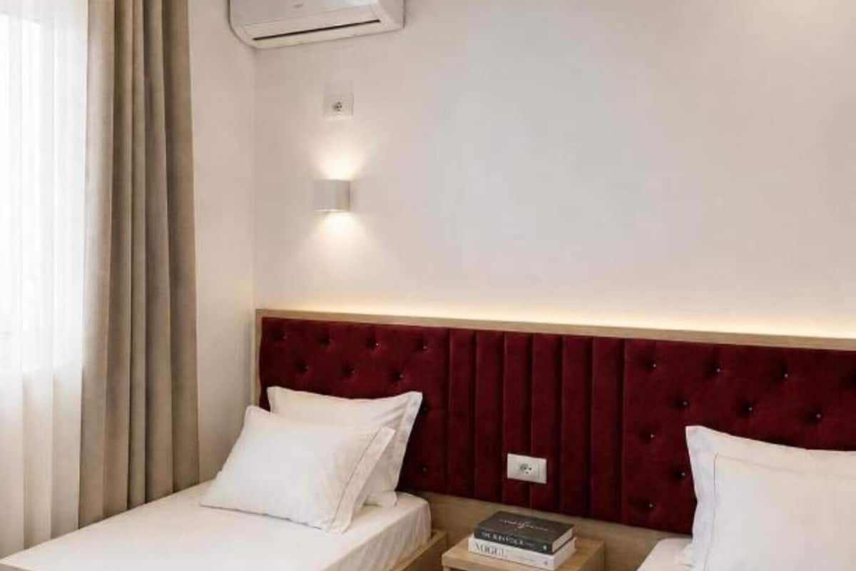 Habitación de hotel con dos camas, cabecero rojo, equipo de aire acondicionado y mesita de noche con libros.