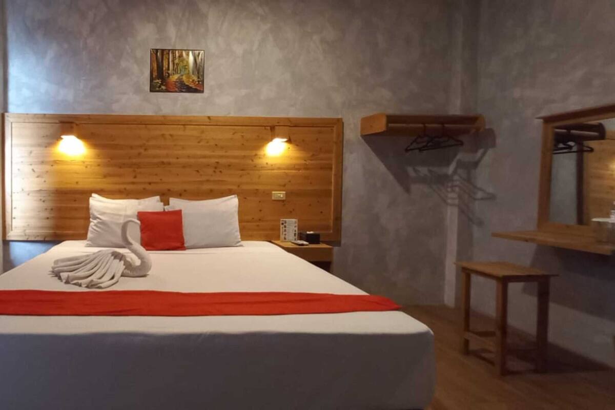 A hotel room with a wooden headboard, a bed with white and red bedding, a red throw blanket, a wooden nightstand, a mirror, and a wooden shelf.