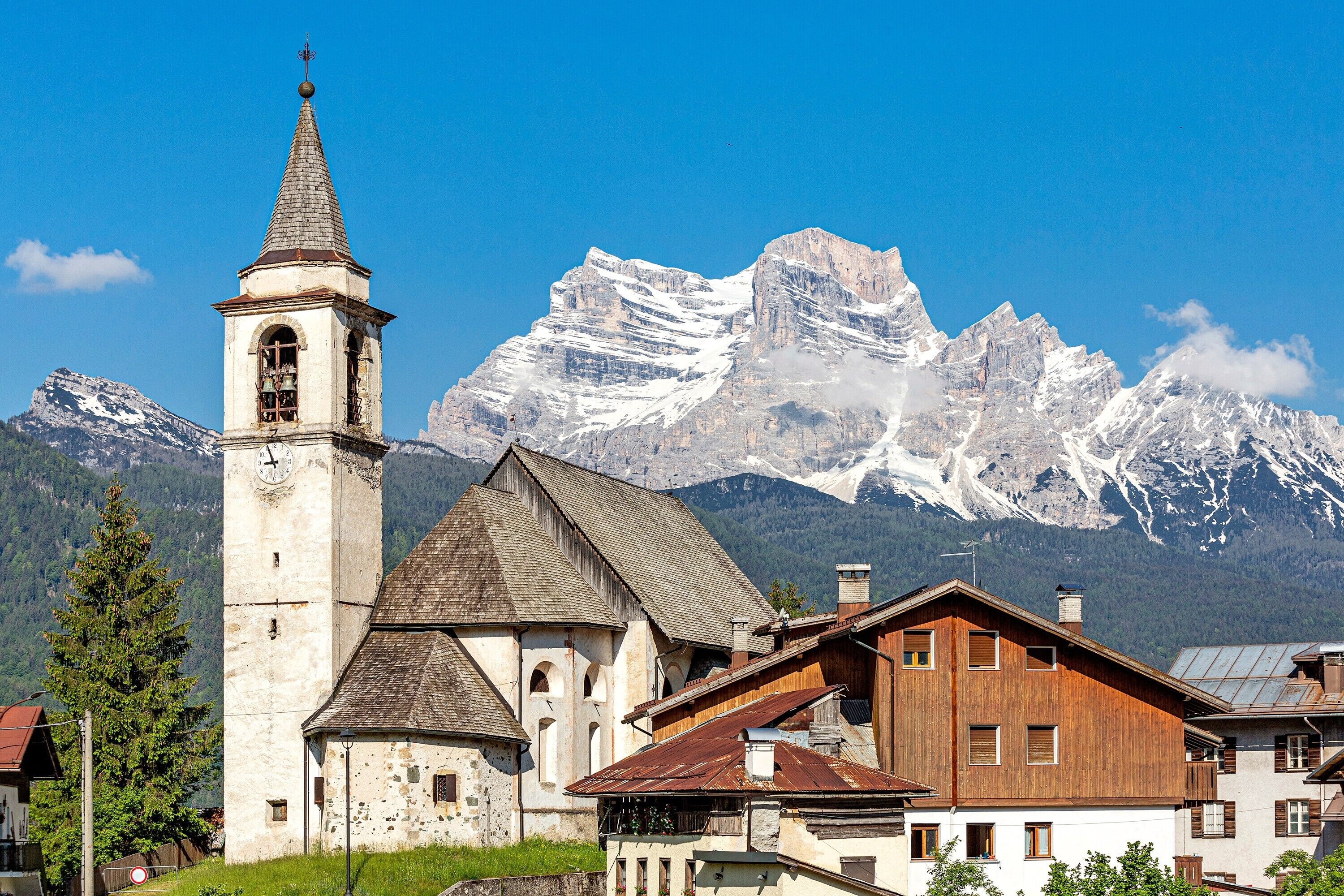 Une église dotée d’un clocher, un bâtiment en bois, et des montagnes enneigées en arrière-plan.