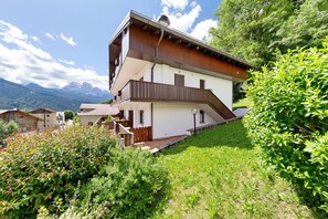 A two-story chalet with a wooden balcony, surrounded by lush greenery and mountains in the background. - Apartment 'Casa Fiori a Vinigo' with Mountain View and Balcony (Vinigo)