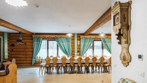 Une pièce aménagée dans le style d’un chalet en rondins, avec une table à manger pour six personnes, des chaises en bois, un canapé à carreaux et une grande horloge murale.