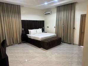 A modern hotel room with a large bed, a dark headboard, beige curtains, and a tiled floor. - SUAVE BY CHLOE (Abuja)
